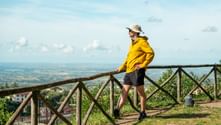 Hiker in yellow jacket and sun hat stands at wooden railing overlooking Italian countryside with expansive valley views under blue sky.