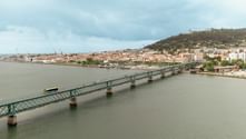 Metal bridge crossing a wide river with a bus on it. Cityscape with orange rooftops and green hills in the background under cloudy skies.