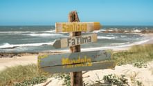 Wooden signpost with arrows pointing to Santiago, Fatima, and Mundele on a sandy beach. Ocean waves and blue sky in background.