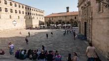 Stenplads i Santiago de Compostela med historisk katedral og bygninger. Grupper af pilgrimme sidder og går over pladsen.
