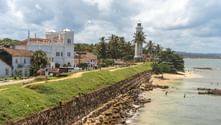 Vit fyr och koloniala byggnader på gräsbevuxna vallar i Galle Fort, Sri Lanka. Stenbefästning med utsikt över strand med palmer.