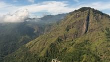 Flygvy över gröna bergstoppar täckta med tät vegetation i Sri Lanka. Moln driver över avlägsna berg under blå himmel.
