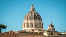 St. Peter's Basilica dome rising above Roman rooftops under clear blue sky. A smaller dome is visible to the right, with terracotta roofs below.