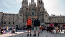 To vandrere med rygsække foran katedralen i Santiago de Compostela. Den gotiske katedral dominerer baggrunden med folkemængder.