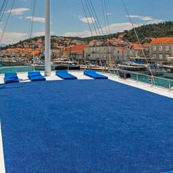 Blåt tæppedækket soldæk på MS Azimut med hvide rækværk, med udsigt over kroatisk kystby med stenbygninger og havn.