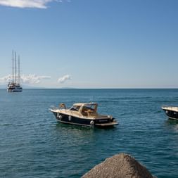 Flere både for anker i turkisblåt Middelhav ved Amalfikysten. To motorbåde i forgrunden, sejlbåde i baggrunden under blå himmel.