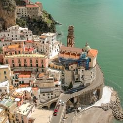 Luftfoto af farverige bygninger der falder ned ad stejle klipper mod turkisblåt Middelhav på Amalfikysten, med kirketårn.