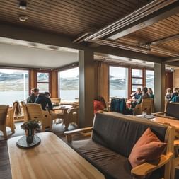 Interiør i DNT Finsehytta hytte med træmøbler, mørke sofaer med orange puder og store vinduer med udsigt over sø og bjerge.