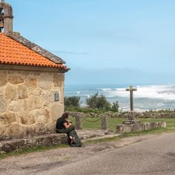 Stone chapel with orange tile roof and bell tower beside coastal road. Two pilgrims rest near the building with ocean waves visible in background.
