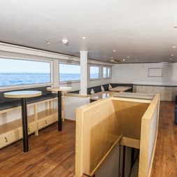 Moderne skibsspisesal med træborde, bænke og store vinduer med havudsigt. Lyse trægulve og hvidt loft.