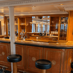 Elegant bar ombord på MS Casanova med buet trædisk, sorte lædertaburetter, udsmykket loft og vinduer med udsigt over vandet.