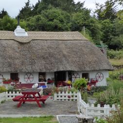Hvidt irsk hus med tykt stråtag og røde vinduesrammer. Rødt picnicbord på stenbelagt terrasse omgivet af hvidt hegn og have.