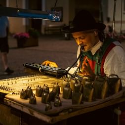 Ældre gademusiker i traditionel vest spiller et stort hackbræt på et bytorv om natten, oplyst af varm gadebelysning.