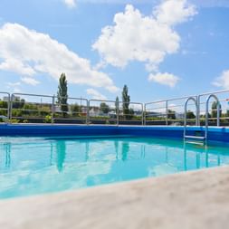 Swimmingpool på MS SE-MANON med klart blåt vand, metalgelænder og stige. Blå himmel med hvide skyer over dækket.