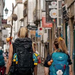 Two pilgrims with backpacks walking through a narrow Portuguese town street. One carries a walking stick with a scallop shell, symbol of the Camino.