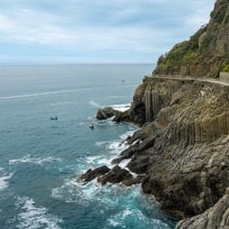 Dramatic coastal path carved into steep rocky cliffs in Cinque Terre, Italy, overlooking turquoise Mediterranean waters with boats visible below.