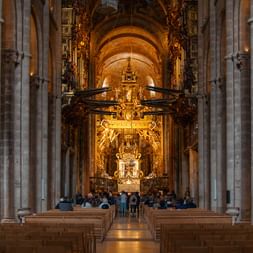 Interior of a cathedral with tall stone columns, wooden pews, and an ornate golden baroque altar illuminated by warm lighting.