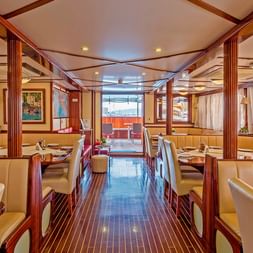 Interiør af MS Amore spisestue med beige læderbænke, træborde, polerede gulve og store vinduer med hvide gardiner.