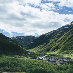 Lille fjeldbebyggelse Tyristhytte i grønne Aurlandsdalen-dal, omgivet af skovklædte skråninger og snedækte bjerge under skyet himmel.