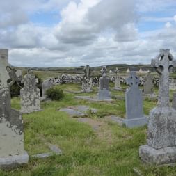 Gammel irsk kirkegård med vejrbidte keltiske stenkors og gravsten spredt over grønt græs, omgivet af stenmure under skyet himmel.