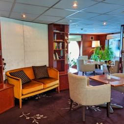 Moderne bibliotek ombord på MS SE-MANON med træbogreolerne, orange sofa, beige lænestole omkring træbord og store vinduer.