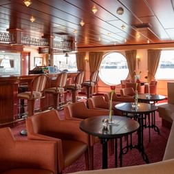 Elegant salonbar på skibet Switzerland med træbar, læderstole, runde borde og runde koøjer med udsigt over vandet.