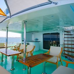 Moderne udendørs spiseplads på High Wave liveaboard med hvide stole, træborde og turkis loft med udsigt over blåt hav.