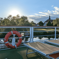 Soldæk på MS Casanova med rød redningskrans, liggestole og hvidt rækværk med udsigt over stille vand. Kirketårn og træer synlige i baggrunden.