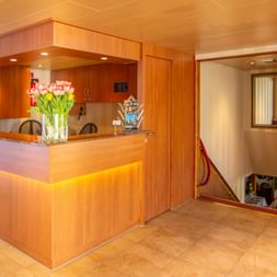 Moderne reception på MS Olympia med trædisk, lyserøde tulipaner i vase og varm belysning. Flisegulv og træpaneler.