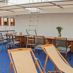 Øverste dæks lounge på MS Azimut med liggestole, spiseborde, træskænk og blåt tæppe. Hvide vægge og orange-indfattede vinduer synlige.