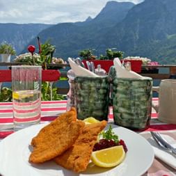 Wienerschnitzel serveret på hvid tallerken med citronskive og tyttebærsauce på ternet dug, bjergudsigt i baggrunden.