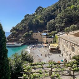 San Fruttuoso beach cove with stone buildings, turquoise water, and forested hillside. Visitors on pebble beach, cypress trees frame the view.