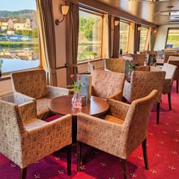 Interiør af MS Olympia skibslounge med beige lænestole omkring små borde, store vinduer med flod- og landskabsudsigt, rødt tæppe.