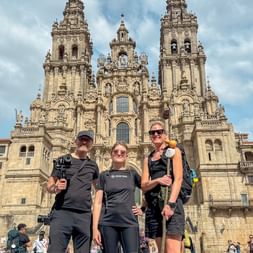 Tre vandrere poserer foran katedralen i Santiago de Compostela på dag 14 af den portugisiske Camino, med barokke tårne bag dem.