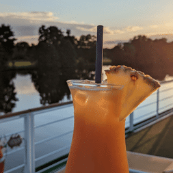 Orange tropisk drink med ananas pynt på MS Casanovas soldæks rækværk ved solnedgang, med rolig flod og træer i baggrunden.