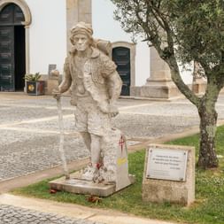 Stone statue of a pilgrim with walking stick and hat on a cobblestone square. Church entrance and olive trees visible in background.