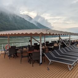 Soldæk på MS Prinzessin Katharina med liggestole, spiseborde under markise og bjerglandskab med skyer i baggrunden.