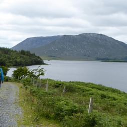 Vandrer med rygsæk går på grusvej langs Western Way-ruten i Irland, med stor sø og bjerge i baggrunden under skyet himmel.