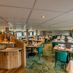 Elegant restaurant ombord på MS Princess med turkise stole, hvide duge og buet træbar. Store vinduer giver naturligt lys.