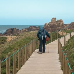 Hiker with backpack and walking poles on wooden boardwalk along Portuguese Camino coastal path, with rocky coastline and ocean view.