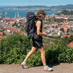 Female hiker with backpack walking on Portuguese Camino Day 10, overlooking coastal town with harbor, residential areas, and mountains in background.