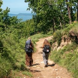 Two hikers with backpacks walking on dirt trail through green landscape on Portuguese Camino. Ocean visible in distance under blue sky.