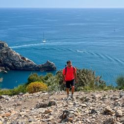 Hiker in red shirt on rocky trail overlooking turquoise Ligurian Sea with historic church on coastal promontory and sailboat in the water.