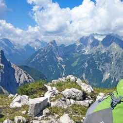 Grøn Vaude rygsæk og vandrestave på klippefyldt top med dramatiske alpetinder i baggrunden. Hvide skyer over Zugspitze bjergkæde.