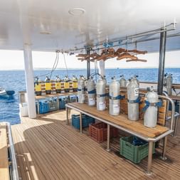 Dykkerdæk på MV Tala med talrige dykkerflasker på træbænke, hængende våddragter og blåt ocean i baggrunden.