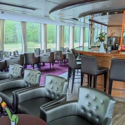 Interiør af MS SE-MANON salon med grå læderlænestole, bar med taburetter, spiseborde og store vinduer med udsigt til grønt landskab.