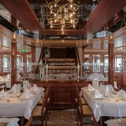 Elegant skibsrestaurant med hvide duge, træstole og historisk skovlhjul som midtpunkt. Spejlloft og varm belysning.