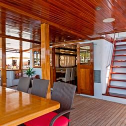 Elegant skibsinteriør med poleret træloft, spisebord med kurvstole, trætrappe og store vinduer med udsigt over vandet.