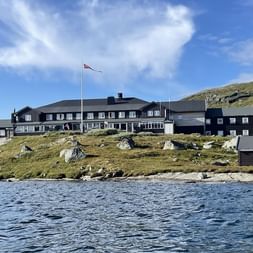 DNT Finsehytta hyttekompleks med mørke træbygninger og norsk flag på klippefyldt terræn ved en bjergsø under blå himmel.