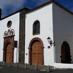 White colonial church with dark volcanic stone trim in Teguise, Lanzarote. Features bell tower, wooden doors, and arched entrance.